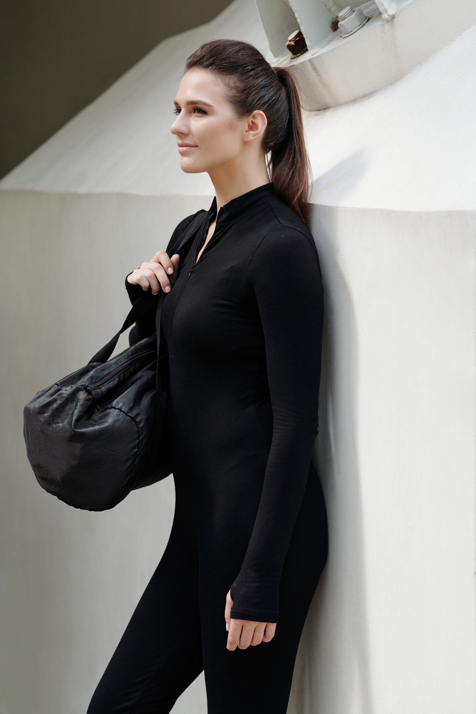 Slim dance girl in black suit standing with sportive bag against white wall outdoors